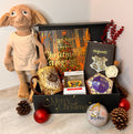 A Harry Potter themed Christmas gift basket arranged in a black box with 'Merry Christmas' written on the front. The basket contains a Christmas in Hogwarts book, a Dobby plush toy, a Marauders Map mug, a Chocolate frog, Harry Potter themed notebook and Christmas ornament and other themed items. Best Christmas Gift for Harry Potter fans.