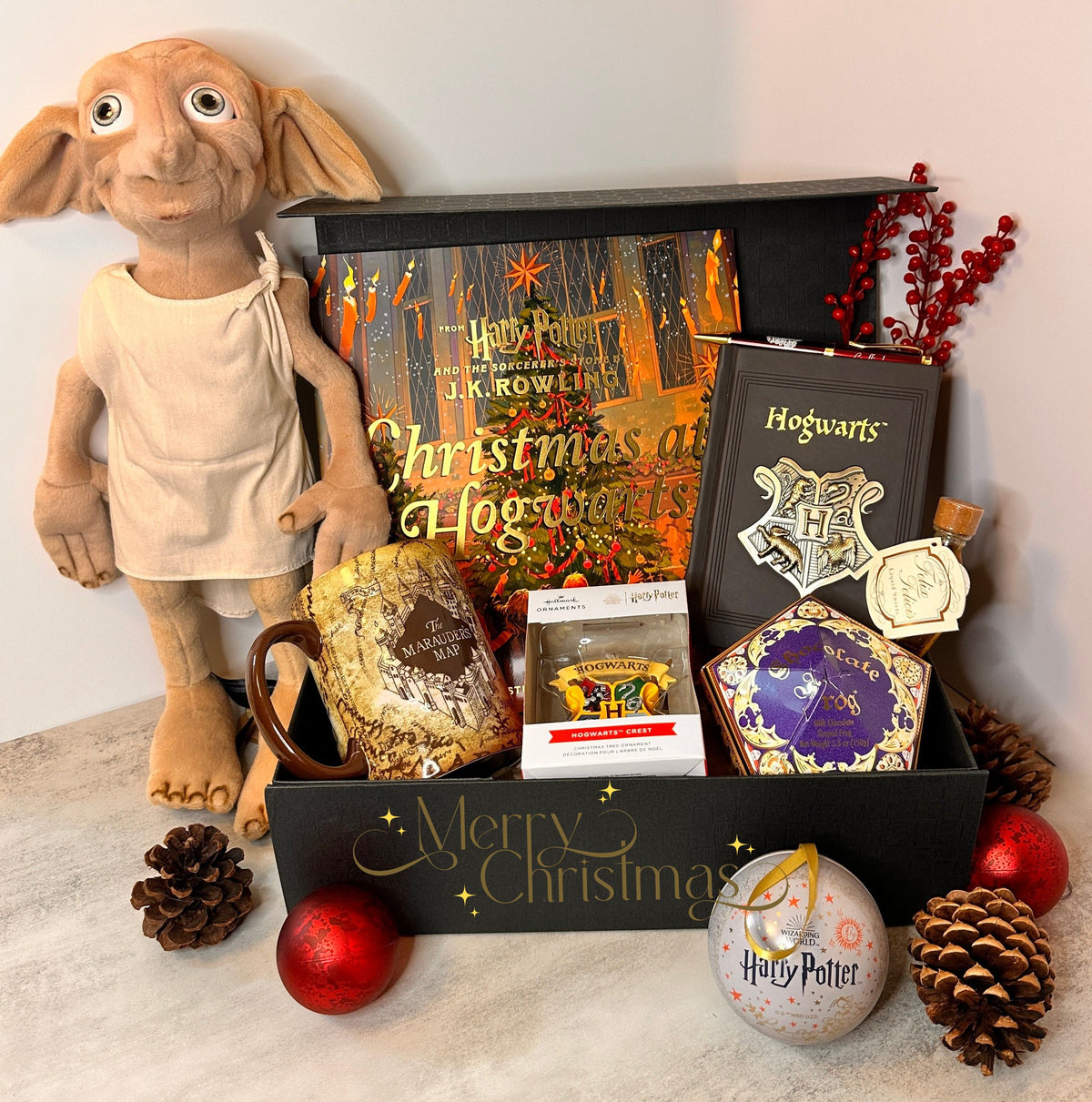 A Harry Potter themed Christmas gift basket arranged in a black box with 'Merry Christmas' written on the front. The basket contains a Christmas in Hogwarts book, a Dobby plush toy, a Marauders Map mug, a Chocolate frog, Harry Potter themed notebook and Christmas ornament and other themed items. Best Christmas Gift for Harry Potter fans.