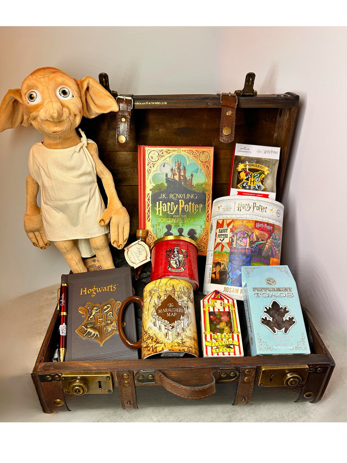 A curated Harry Potter themed gift basket arranged inside an open vintage-style  Harry Potter brown suitcase. Visible items include a Dobby plush figure, a Hogwarts notebook, a Harry Potter book, a Harry Potter themed candle, Harry Potter themed puzzle, Harry Potter Marauders Map mug , Harry Potter Christmas ornament, Harry Potter sweet treats (chocolate frogs, Felix Felicis Liquid Betrtie Bott's)  