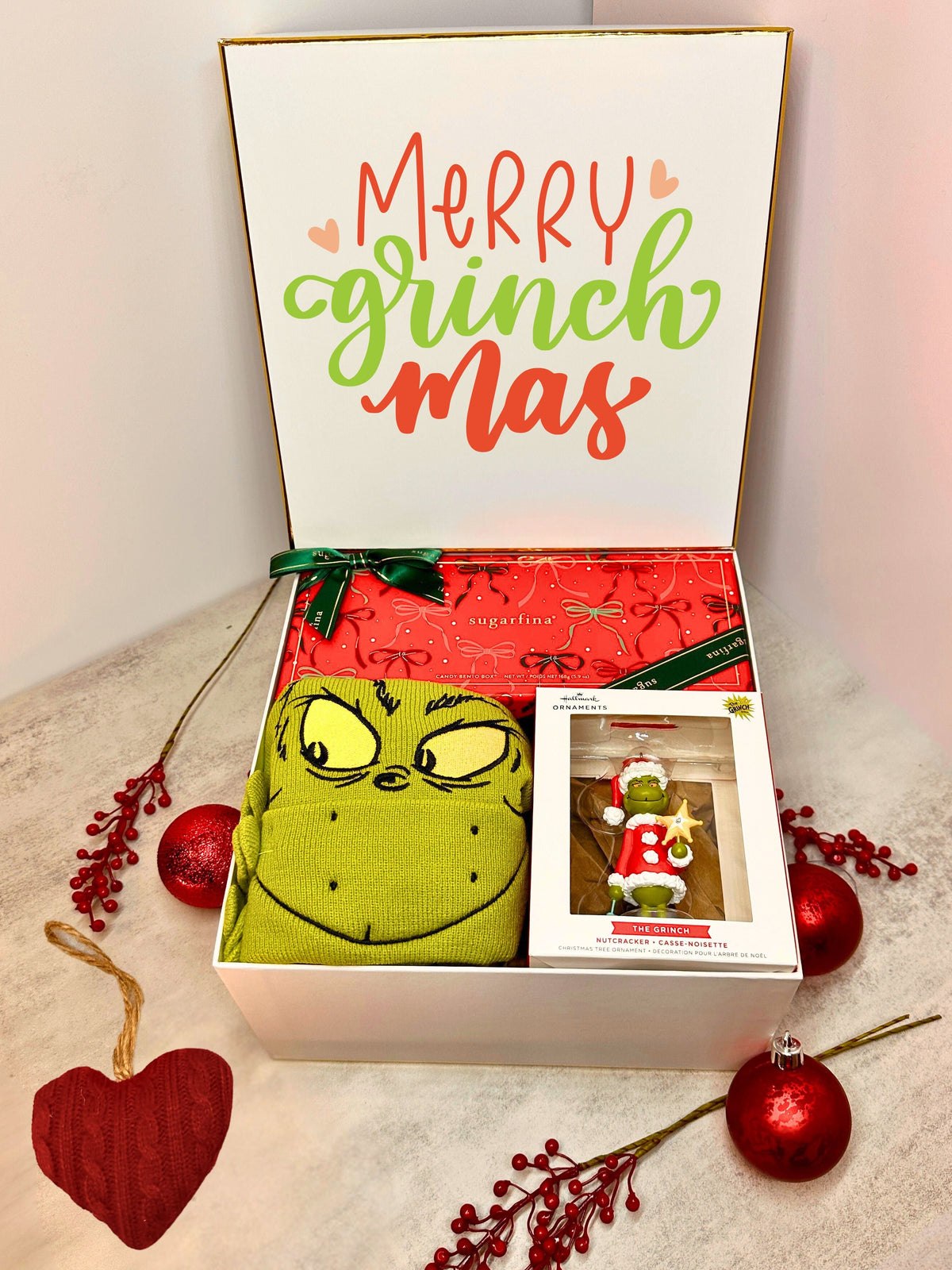 A Christmas gift set in a white box with the lid printed 'Merry grinch mas' in red and green lettering. Inside are a green Grinch-themed hat, Grinch ornament and a Sugarfina candy bento box.