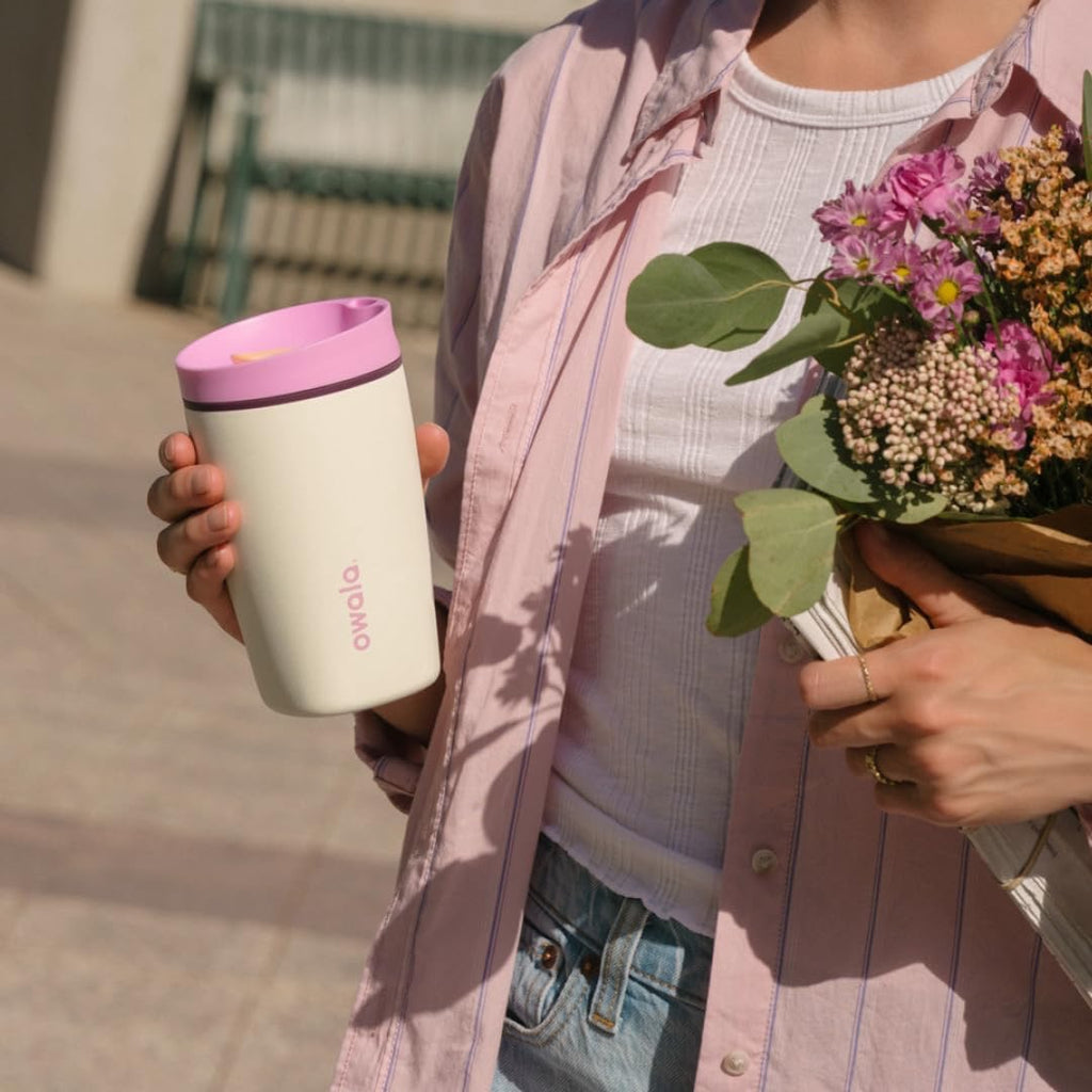 Person holding a pink and white tumbler and a bouquet of flowers outdoors.