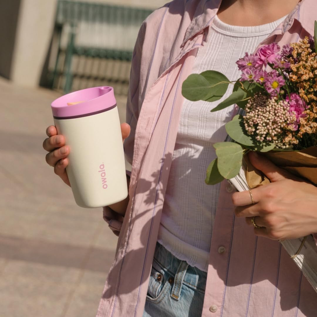 Person holding a pink and white tumbler and a bouquet of flowers outdoors.