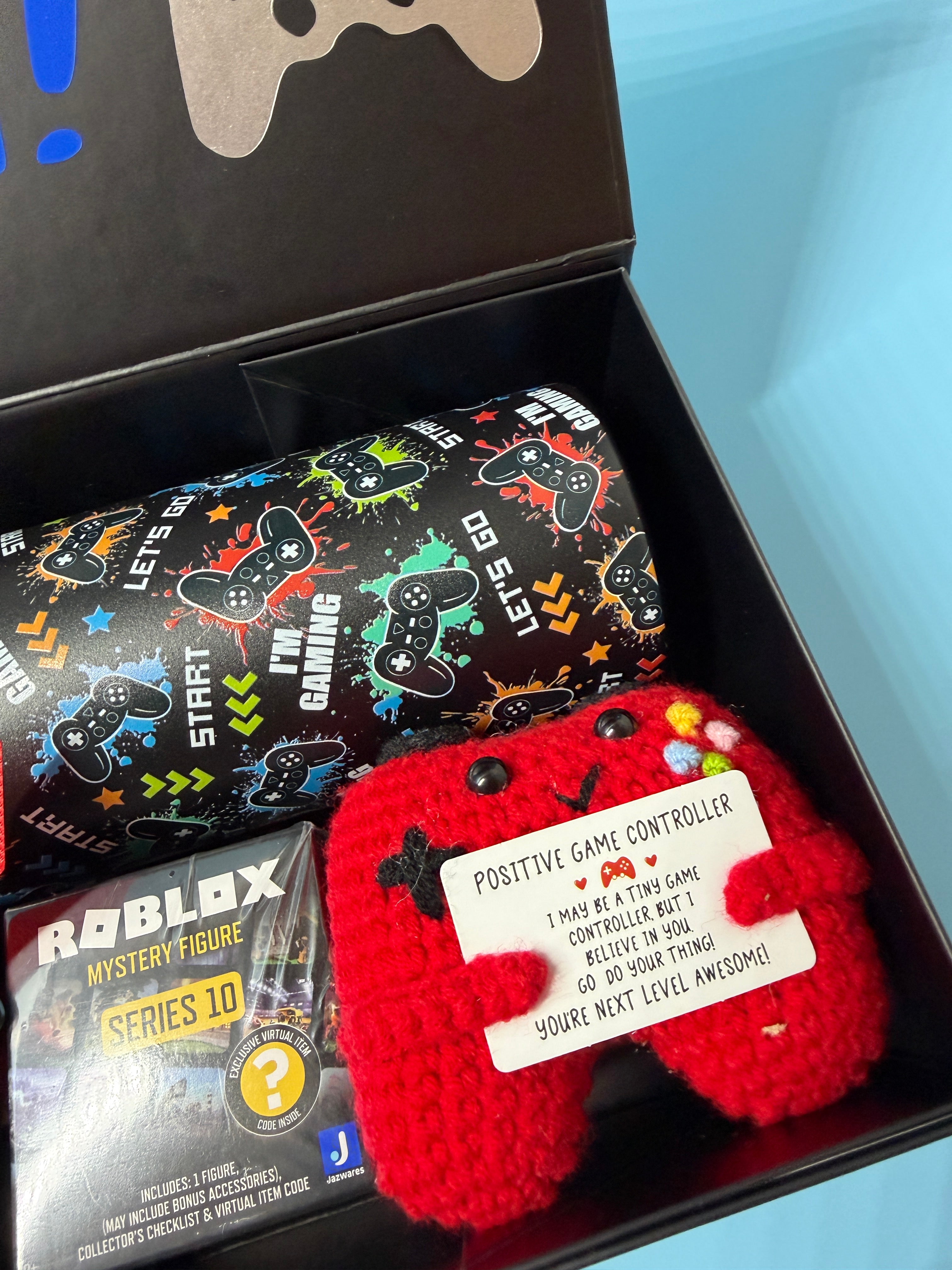 Colorful gaming-themed box with a Roblox collectable figure and a red controller-shaped crochet toy in a box with a blue background.